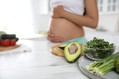 Photo of Fresh vegetables at table and young pregnant woman in kitchen, closeup. Taking care of baby health
