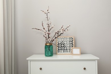 Photo of Flowering tree twigs and decor on white chest of drawers near light wall in room