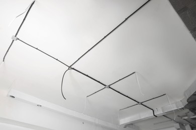 Photo of Conduits with cables and ventilation system on white ceiling, low angle view. Installation of electrical wiring
