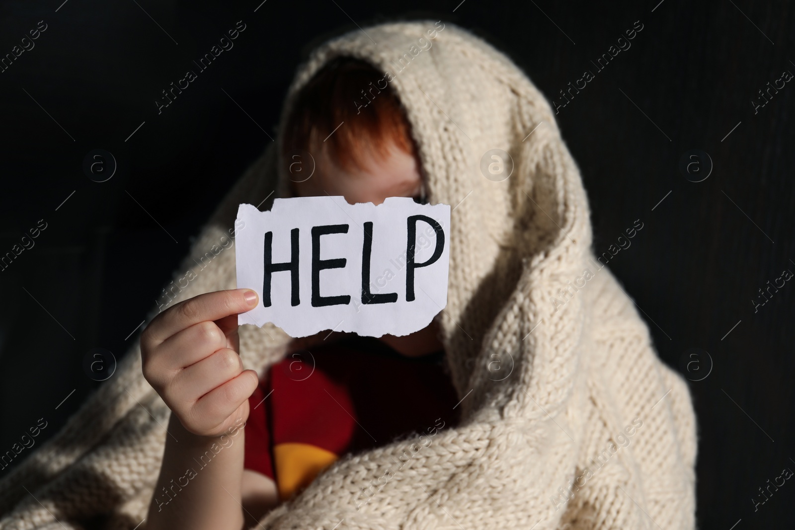 Photo of Little boy wrapped in blanket holding piece of paper with word Help in darkness, focus on hand. Domestic violence concept