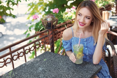 Photo of Young woman with glass of tasty lemonade at table in cafe, outdoors. Natural detox drink