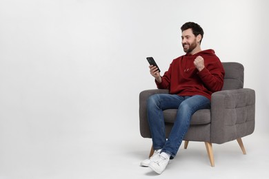 Happy man with smartphone sitting on armchair against white background. Space for text