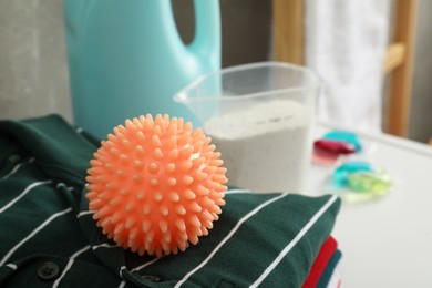 Photo of Orange dryer ball and stacked clean clothes near laundry detergents on white table. Space for text
