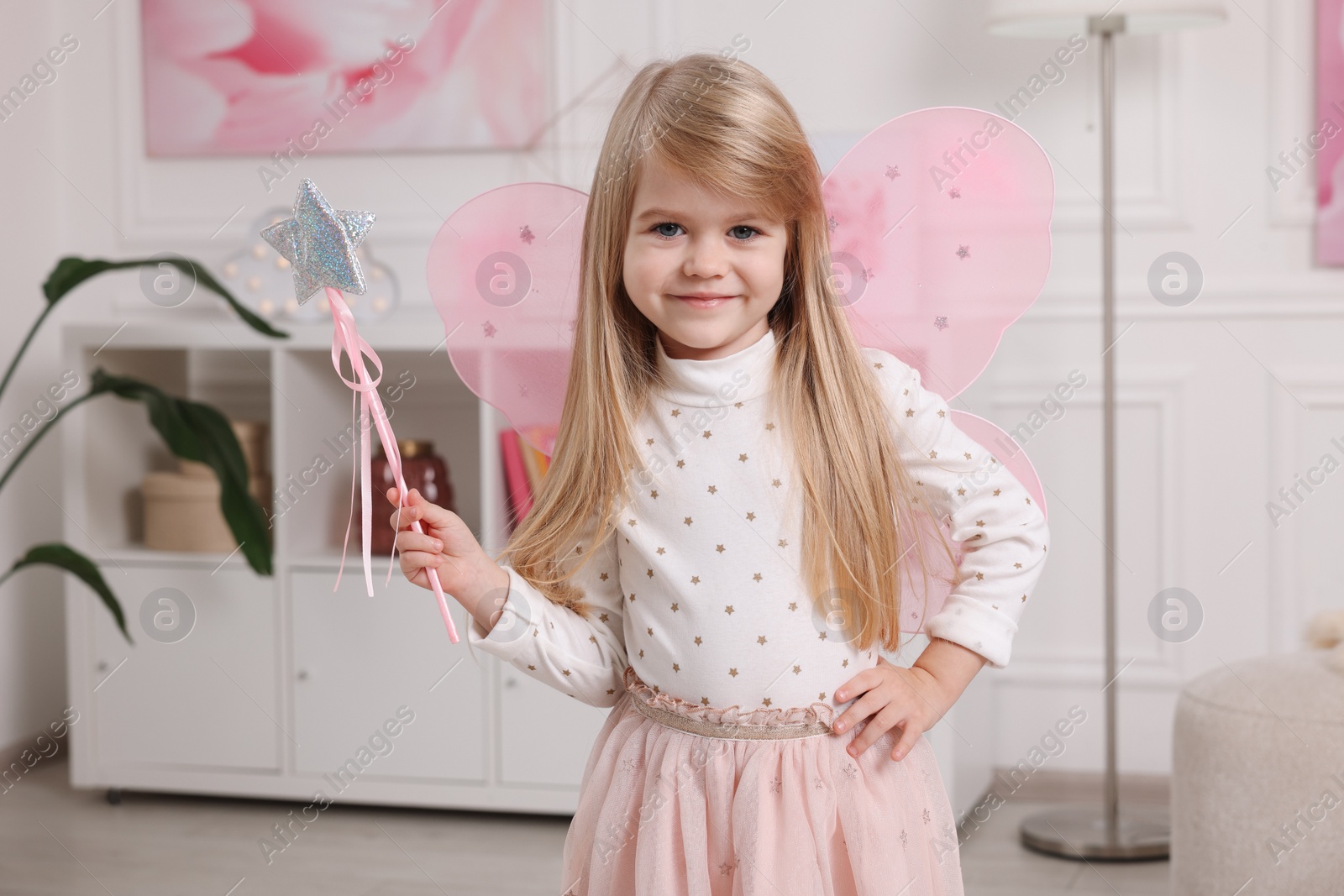 Photo of Cute little girl in fairy costume with pink wings and magic wand at home