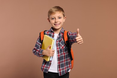 Photo of Happy schoolboy with backpack and books showing thumb up gesture on brown background