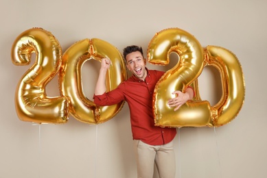 Excited young man with golden 2020 balloons on beige background. New Year celebration