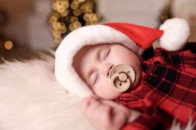 Cute baby in Santa hat with pacifier sleeping on soft faux fur indoors. Christmas season
