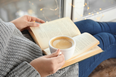 Woman with cup of coffee reading book at home, closeup