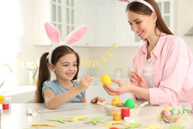 Happy mother and daughter with bunny ears headbands painting Easter egg in kitchen