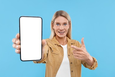 Happy woman holding smartphone and pointing at blank screen on light blue background