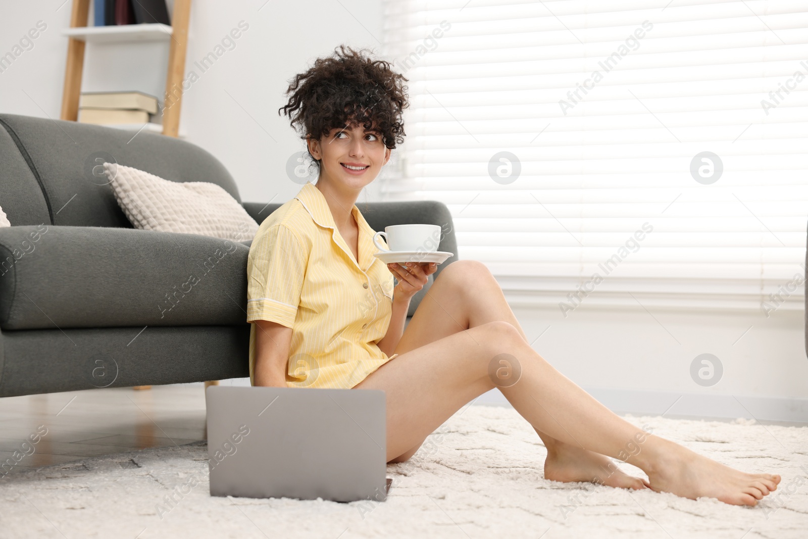 Photo of Beautiful young woman in stylish pyjama with cup of drink using laptop on floor at home