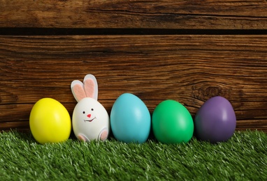 Bright eggs and white one as Easter bunny on green grass against wooden background