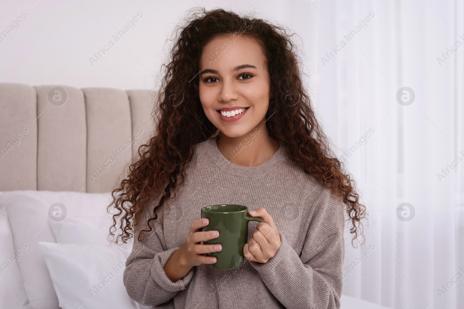 Photo of Happy African American woman with cup of drink in bedroom