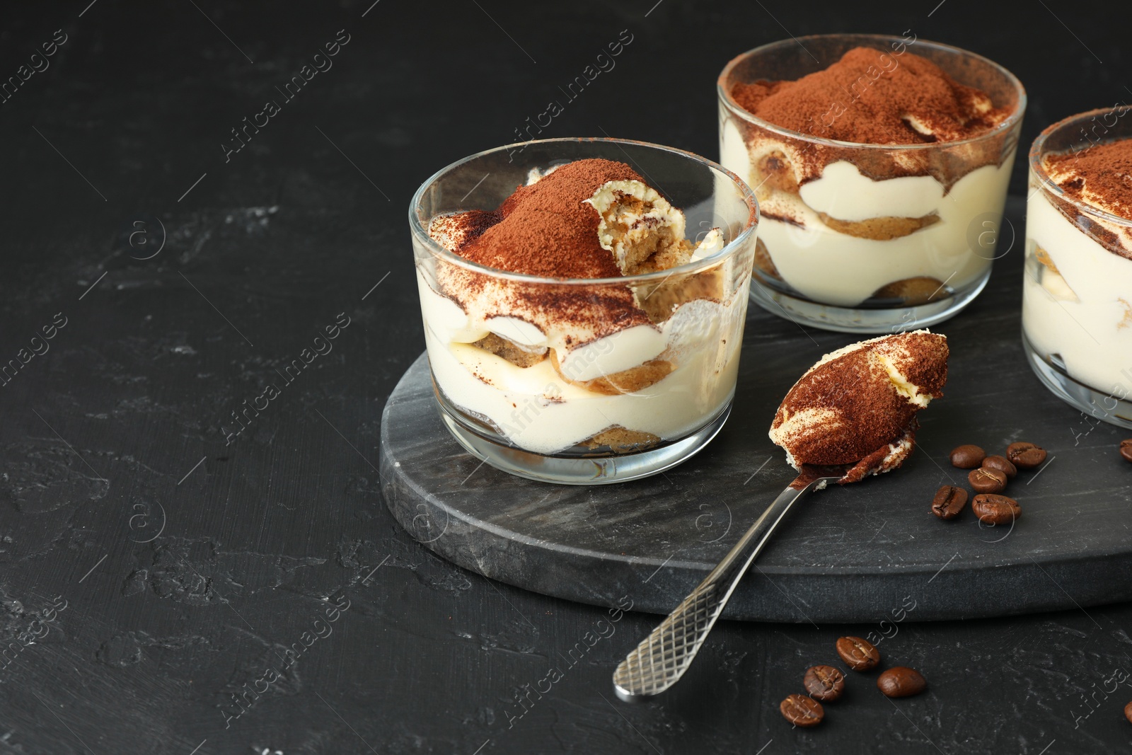 Photo of Delicious tiramisu in glasses, spoon and coffee beans on black table, space for text