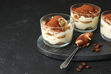 Photo of Delicious tiramisu in glasses, spoon and coffee beans on black table, space for text