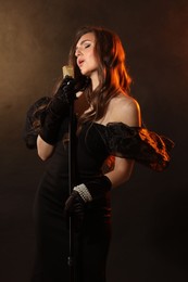 Photo of Beautiful young woman with microphone singing on dark background with smoke