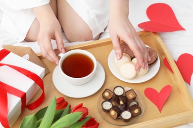 Photo of Tasty breakfast served in bed. Woman with tea, desserts, gift box and flowers at home, closeup