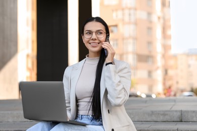 Photo of Happy young woman using modern laptop and talking on smartphone outdoors
