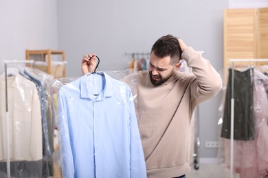 Photo of Dry-cleaning service. Emotional man holding hanger with shirt in plastic bag indoors