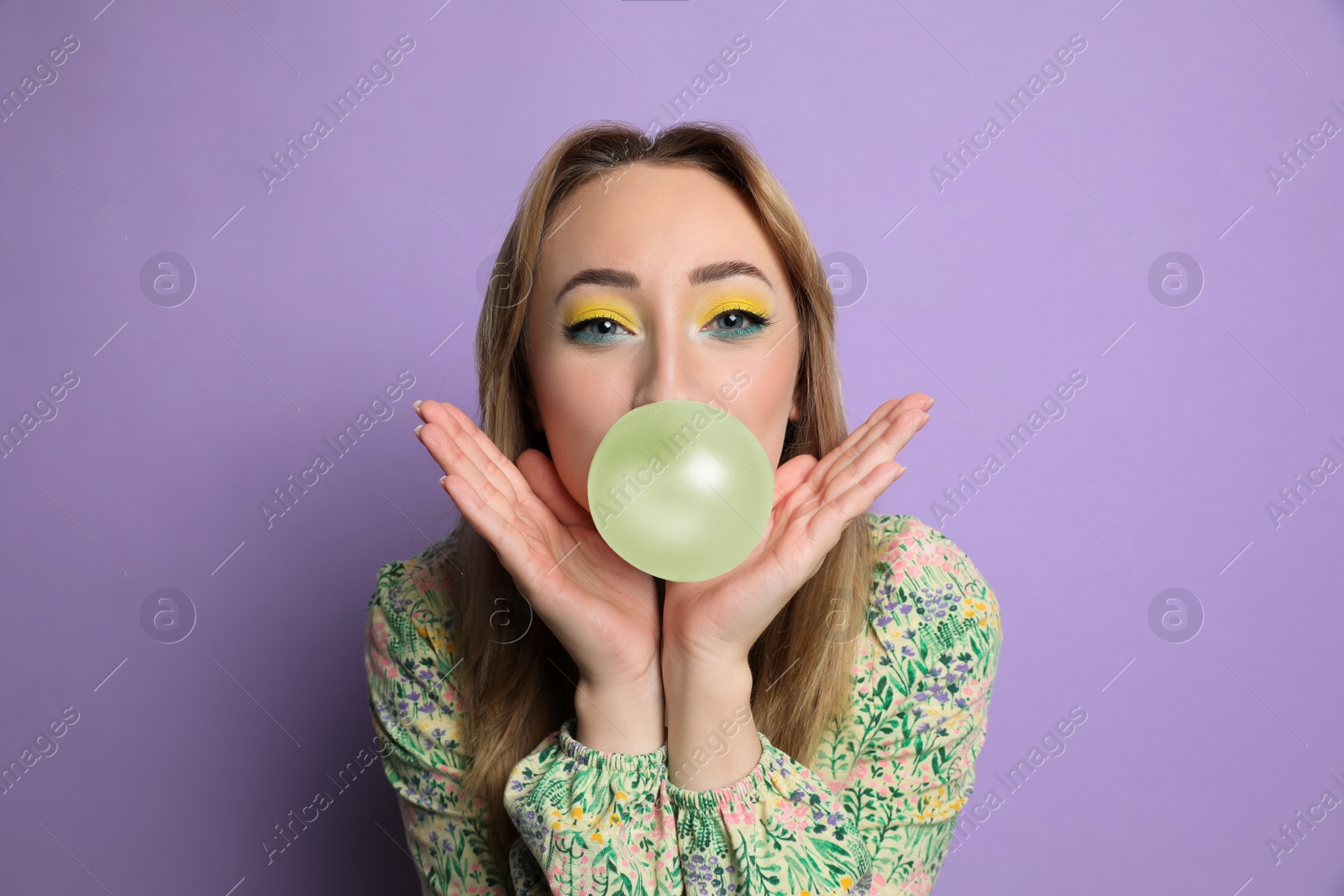 Photo of Fashionable young woman with bright makeup blowing bubblegum on lilac background