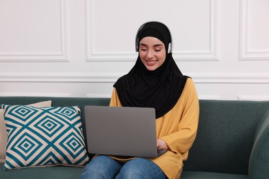 Photo of Muslim woman in hijab and headphones using laptop on sofa indoors