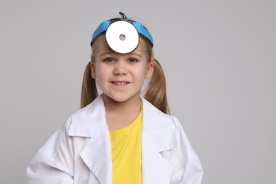 Photo of Little girl in medical uniform with head mirror on light grey background. Space for text