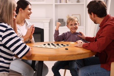 Photo of Family playing checkers at wooden table in room. Son enjoying winning