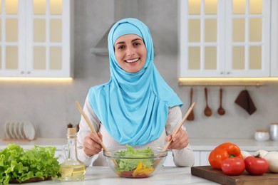 Photo of Muslim woman making delicious salad with vegetables at white table in kitchen