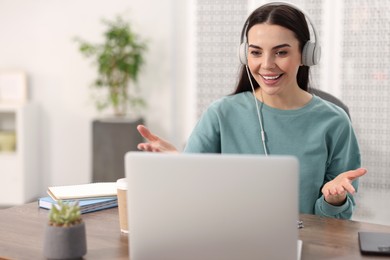 Photo of Young woman in headphones using video chat during webinar at table in room