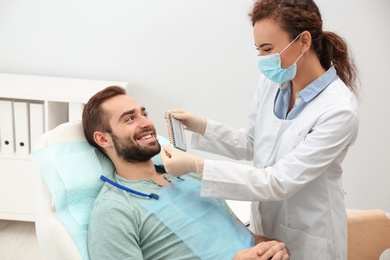 Dentist matching young man's teeth color with palette in office