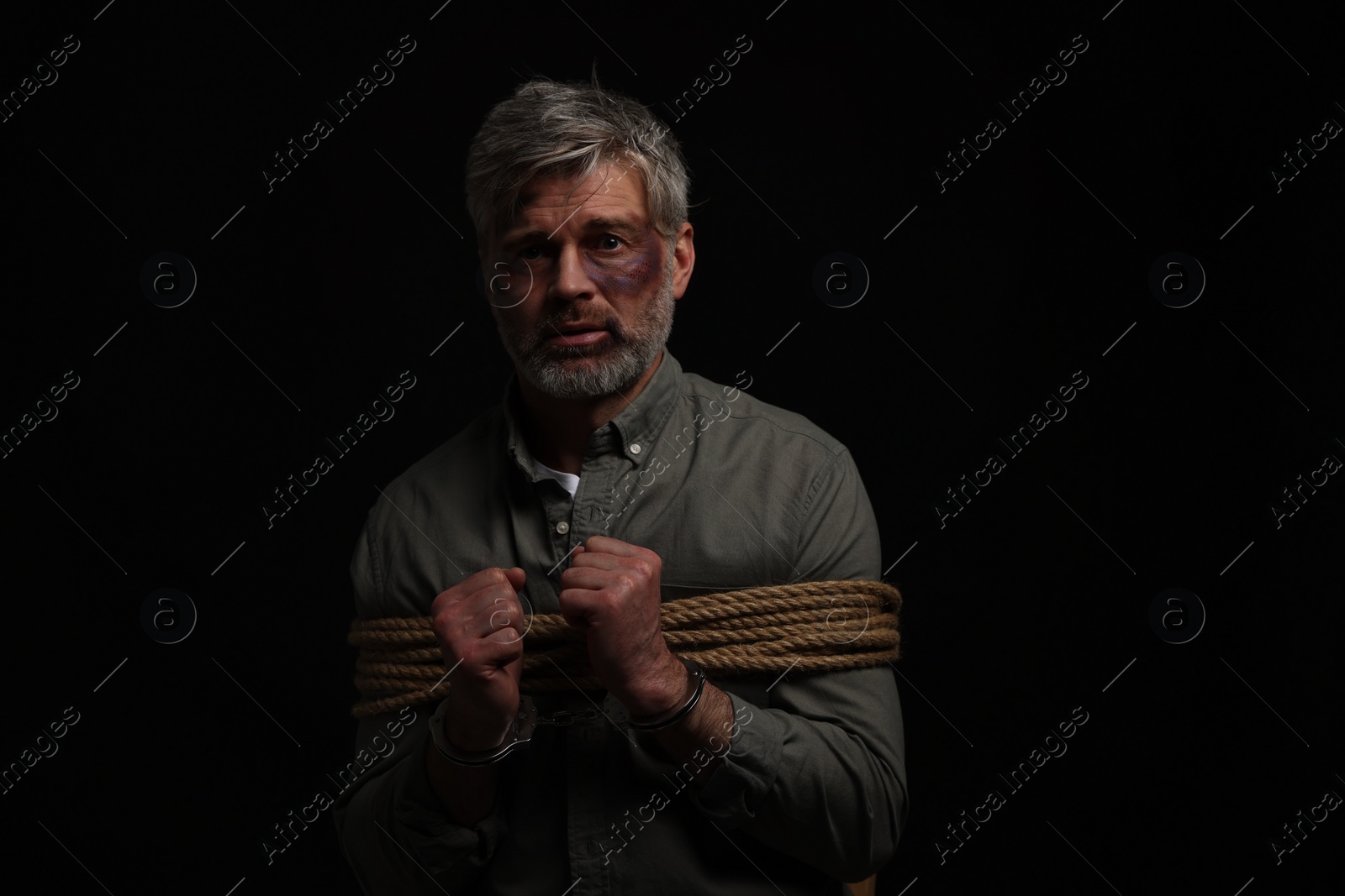 Photo of Beaten man tied with rope and handcuffs on black background. Hostage taking