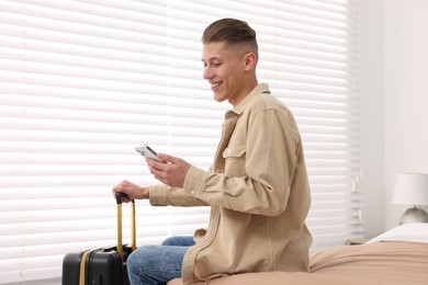 Photo of Smiling guest with suitcase and smartphone on bed in stylish hotel room