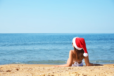 Photo of Young woman wearing Santa hat and bikini on beach, space for text. Christmas vacation