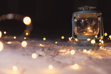 Photo of Lantern with burning candle and Christmas lights on white snow outdoors. Space for text