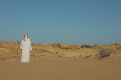 Photo of Man in arabic clothes walking through desert on sunny day, back view