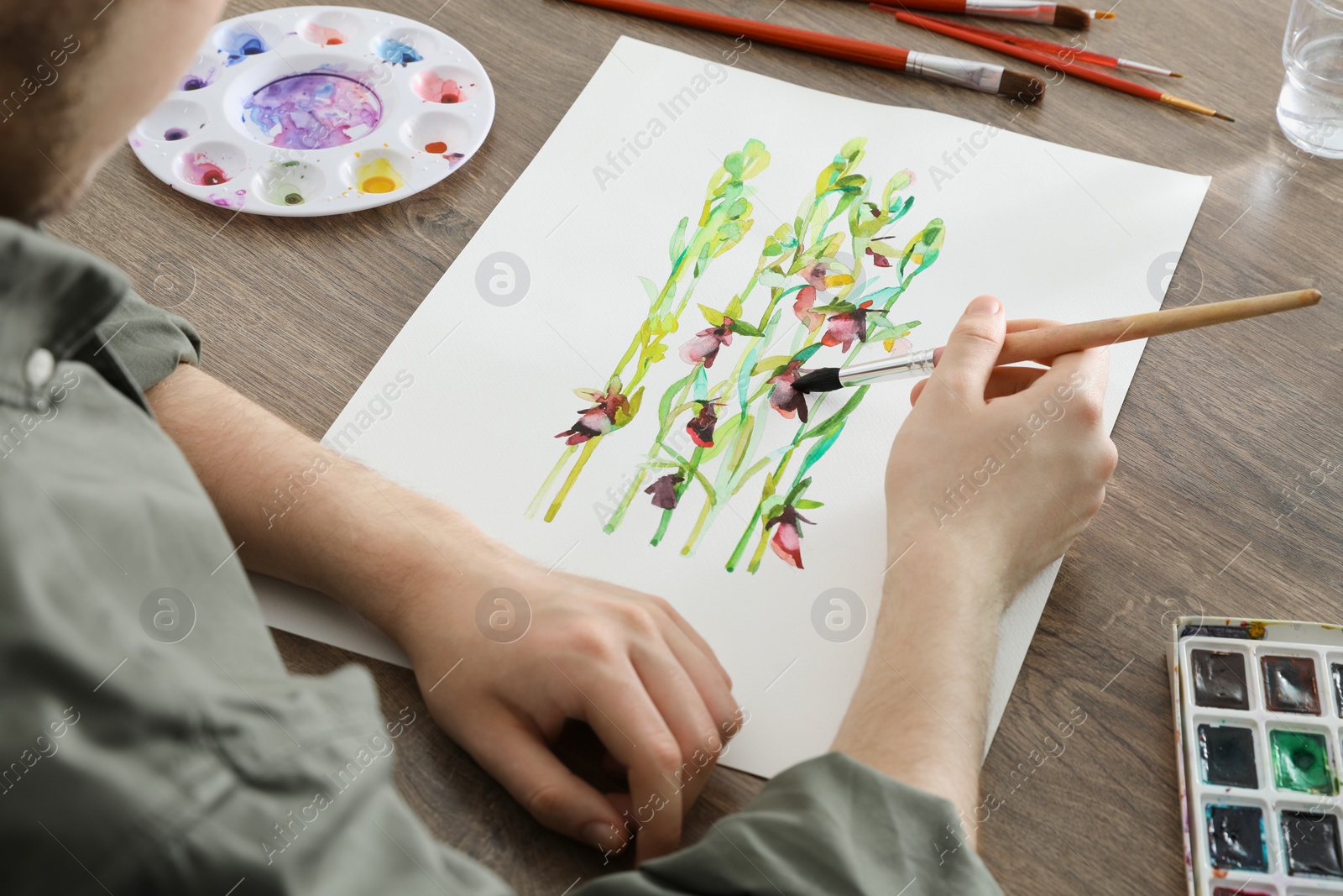 Photo of Man painting flowers with watercolor at wooden table, closeup. Creative artwork