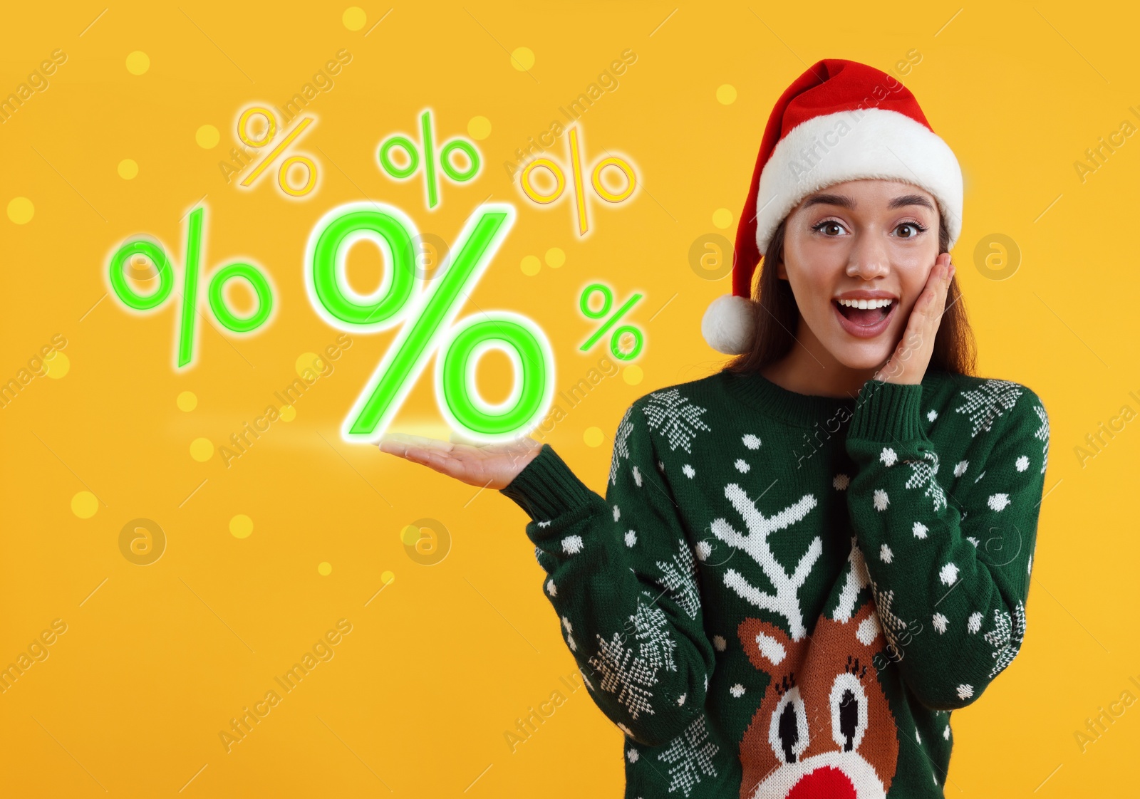 Image of Discount offer. Happy young woman in Christmas sweater and Santa hat showing percent signs on orange background