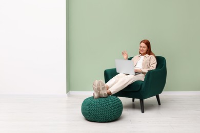 Happy young woman having video chat via laptop while sitting in armchair near light olive wall indoors. Space for text