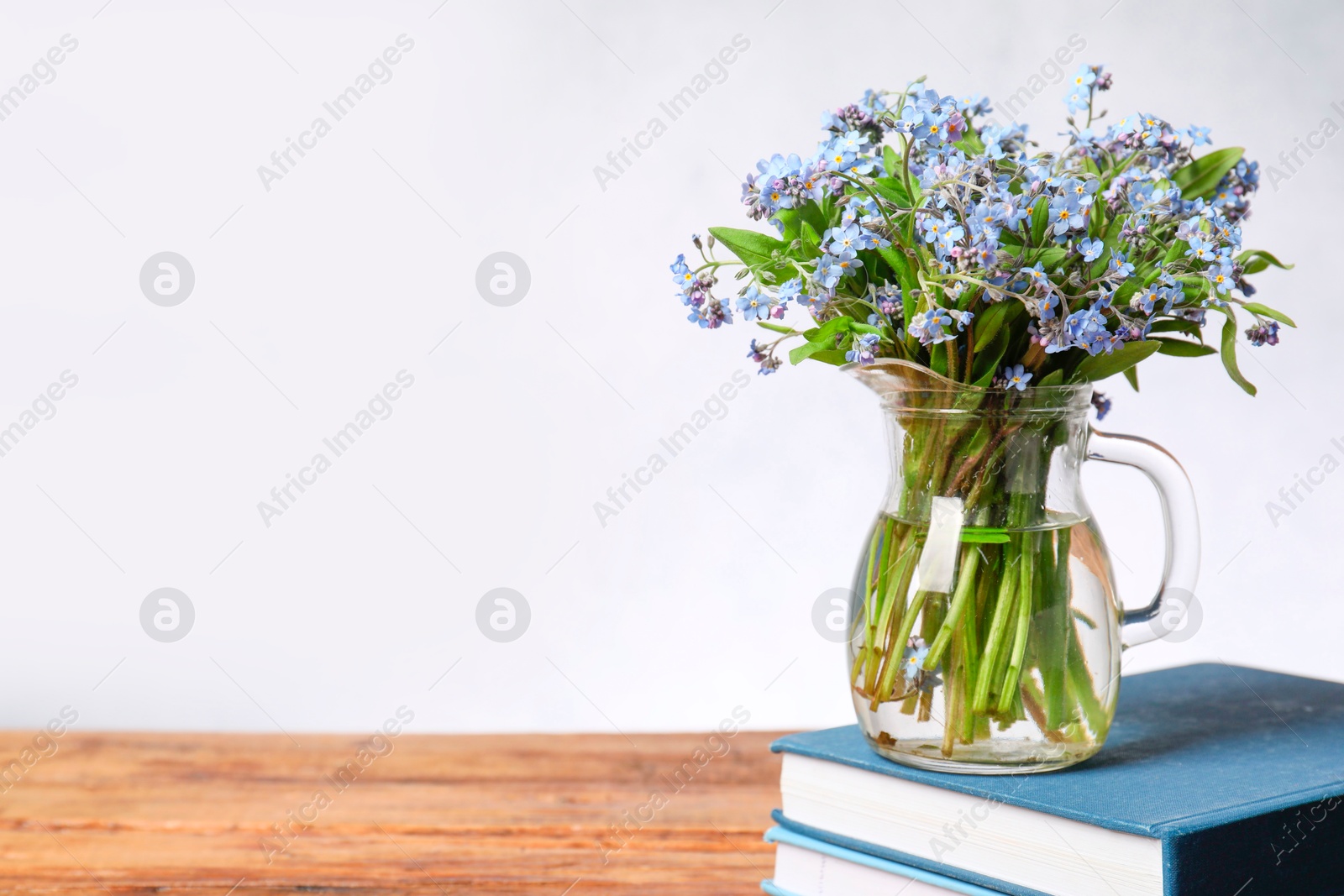 Photo of Bouquet of beautiful forget-me-not flowers in glass jug and books on wooden table against light background, space for text