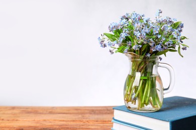 Photo of Bouquet of beautiful forget-me-not flowers in glass jug and books on wooden table against light background, space for text