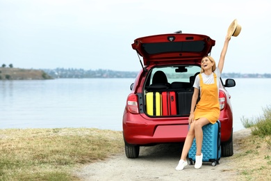 Happy woman near car trunk with suitcases on riverside. Space for text