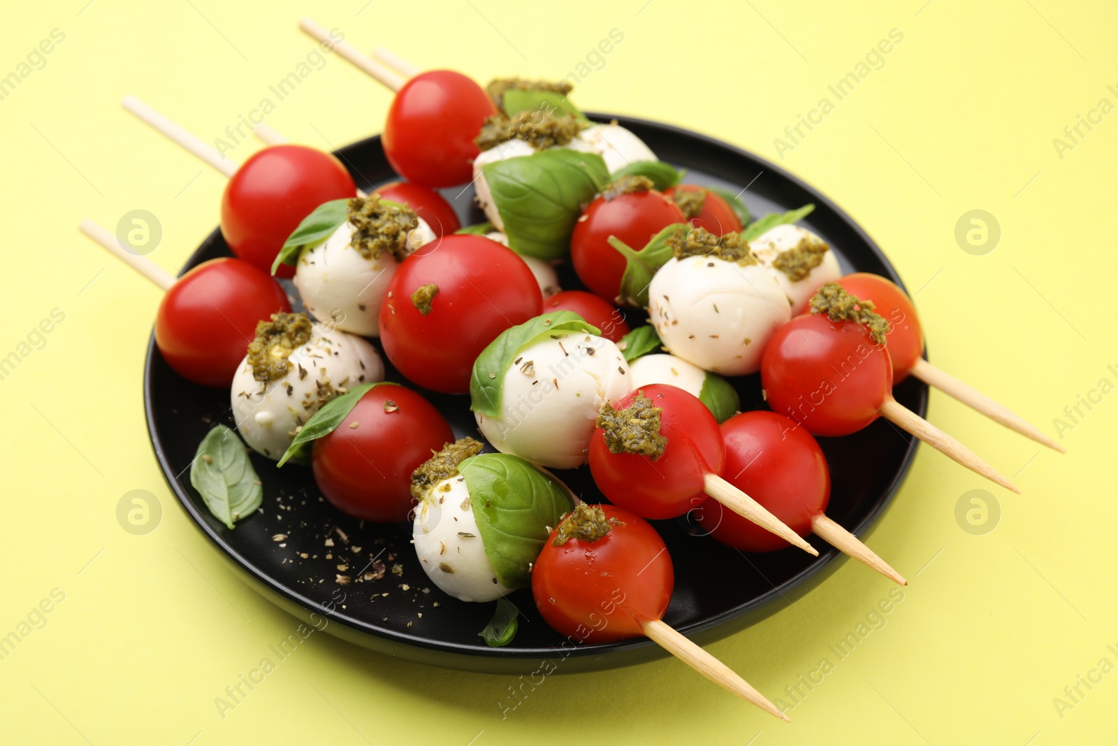Photo of Caprese skewers with tomatoes, mozzarella balls, basil and pesto sauce on yellow background, closeup