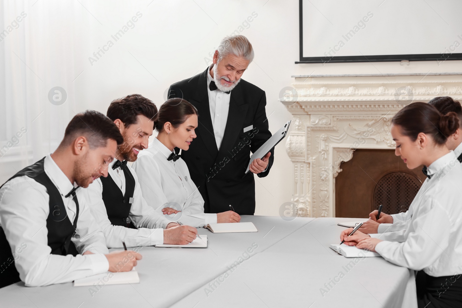 Photo of Senior man in formal suit teaching trainees indoors. Professional butler courses