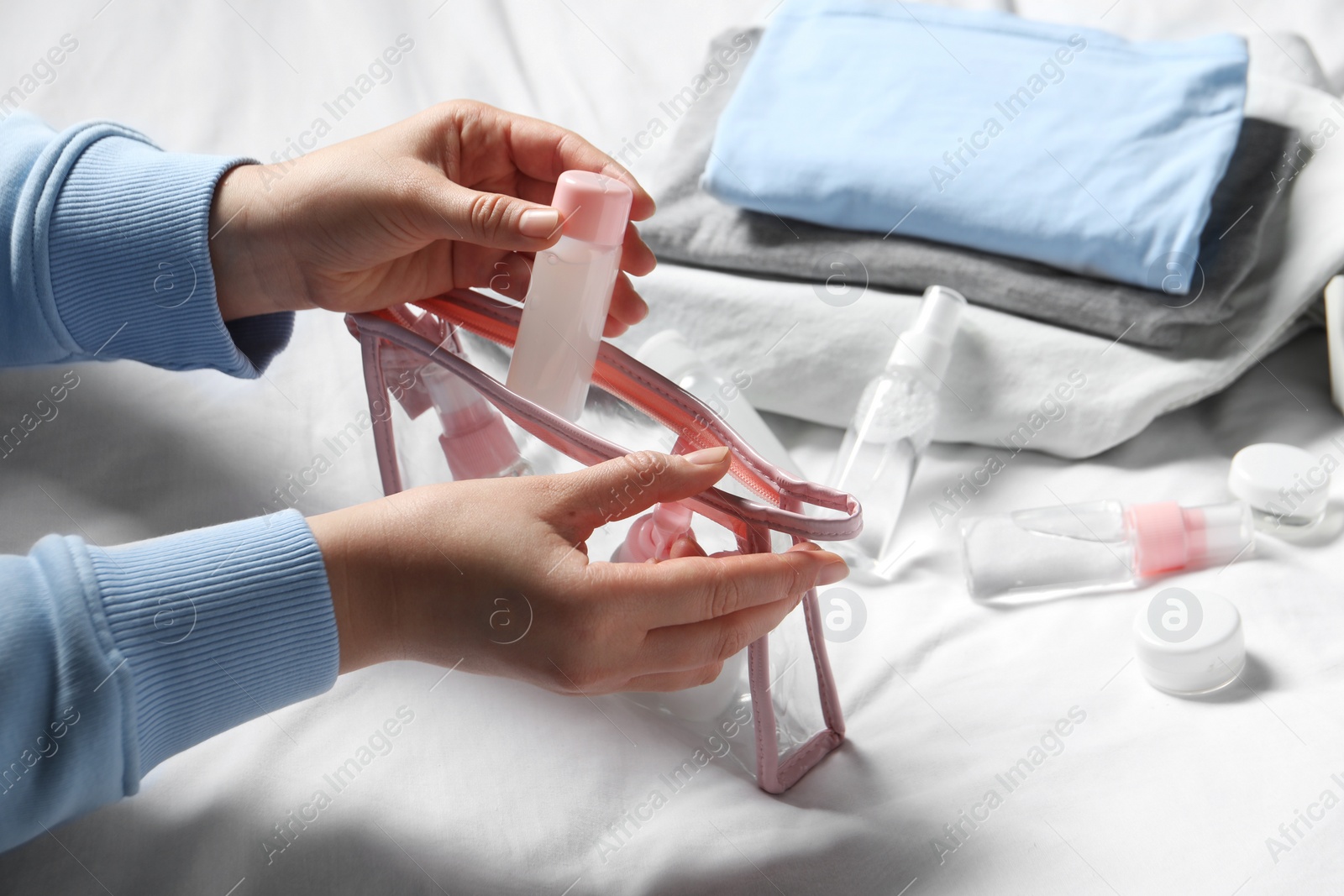 Photo of Cosmetic travel kit. Woman putting small bottle with personal care product into plastic bag on bed, closeup