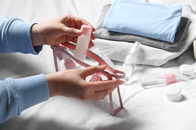 Photo of Cosmetic travel kit. Woman putting small bottle with personal care product into plastic bag on bed, closeup