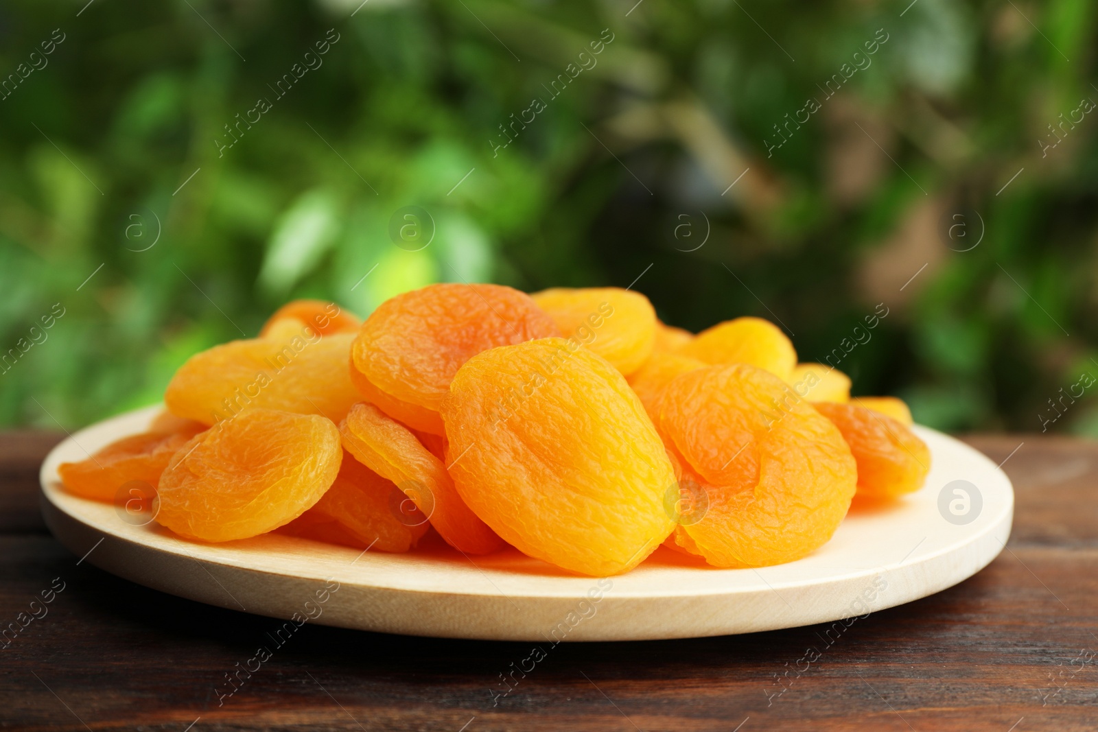 Photo of Plate of tasty apricots on wooden table against blurred green background. Dried fruits