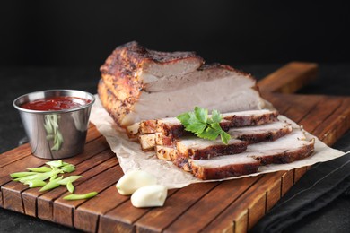 Photo of Pieces of baked pork belly served with sauce and parsley on black textured table, closeup