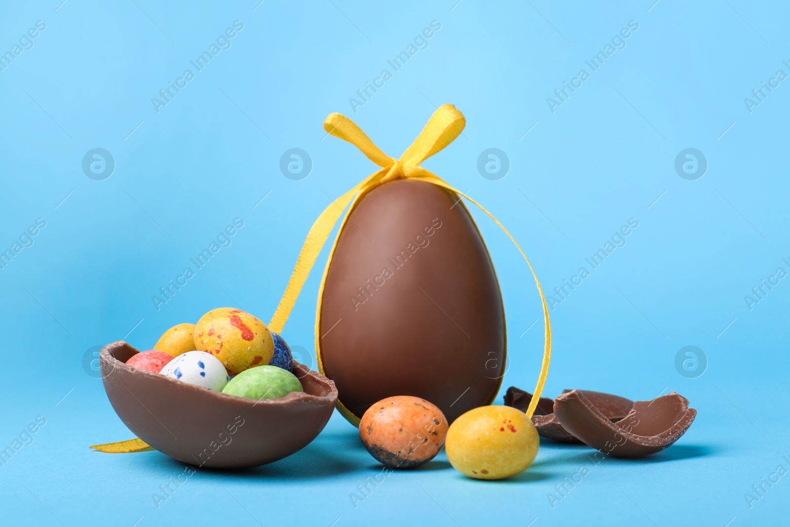 Photo of Tasty whole chocolate eggs with yellow bow and different candies on light blue background