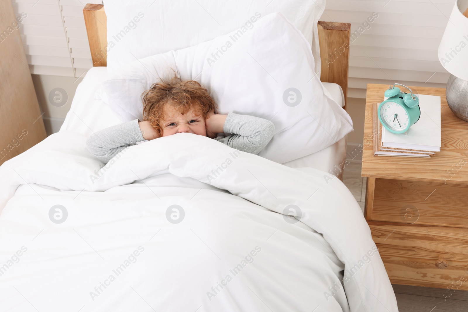 Photo of Angry boy covering ears with hands near alarm clock at early morning indoors