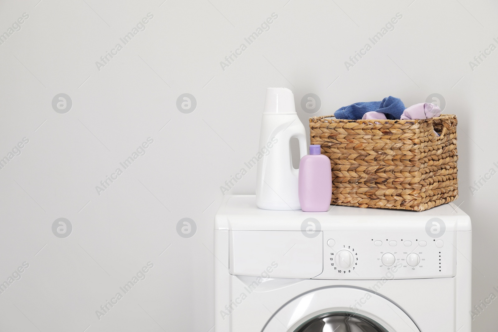 Photo of Baby clothes in wicker basket and laundry detergents on washing machine near light wall, space for text